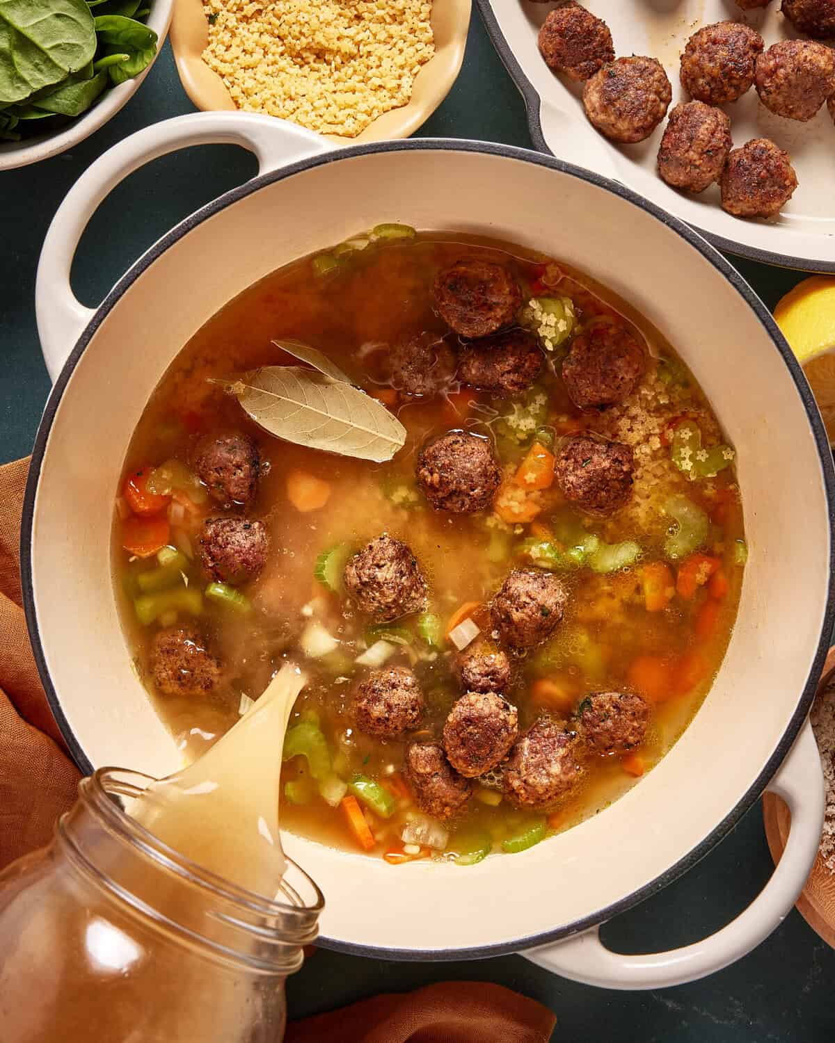 A large pot filled with broth, vegetables, meatballs, and a bay leaf. Broth is being poured in, and bowls of couscous, spinach, and more meatballs are visible nearby on the dark countertop.