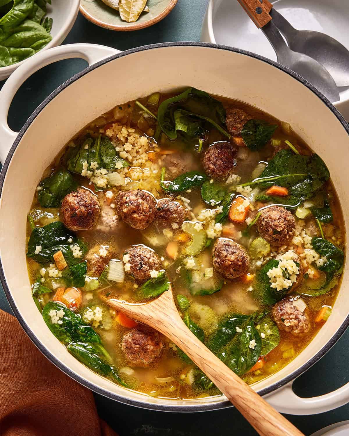 A pot of Italian wedding soup with meatballs, spinach, carrots, celery, pasta, and broth. A wooden spoon rests in the soup, and plates and utensils are visible in the background.