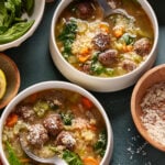 Two bowls of Italian wedding soup with meatballs, spinach, carrots, celery, and pasta, topped with grated cheese. Spoons rest in the bowls, surrounded by bowls of spinach and shredded cheese on the table.