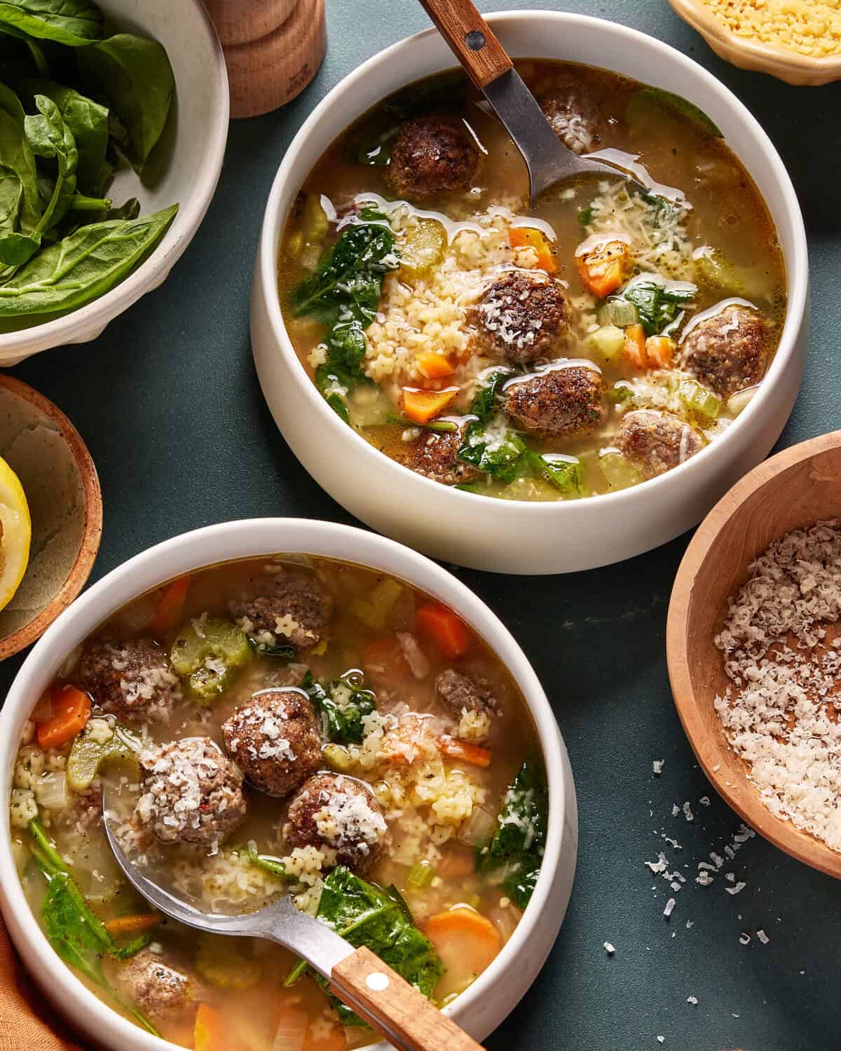 Two bowls of Italian wedding soup with meatballs, spinach, carrots, celery, and pasta, topped with grated cheese. Spoons rest in the bowls, surrounded by bowls of spinach and shredded cheese on the table.