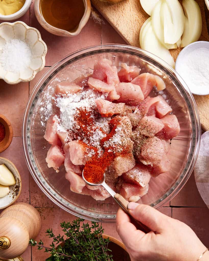 A hand sprinkles paprika over a bowl of raw, diced chicken, surrounded by bowls of salt, spices, garlic, oil, onion slices, and fresh herbs on a kitchen counter.