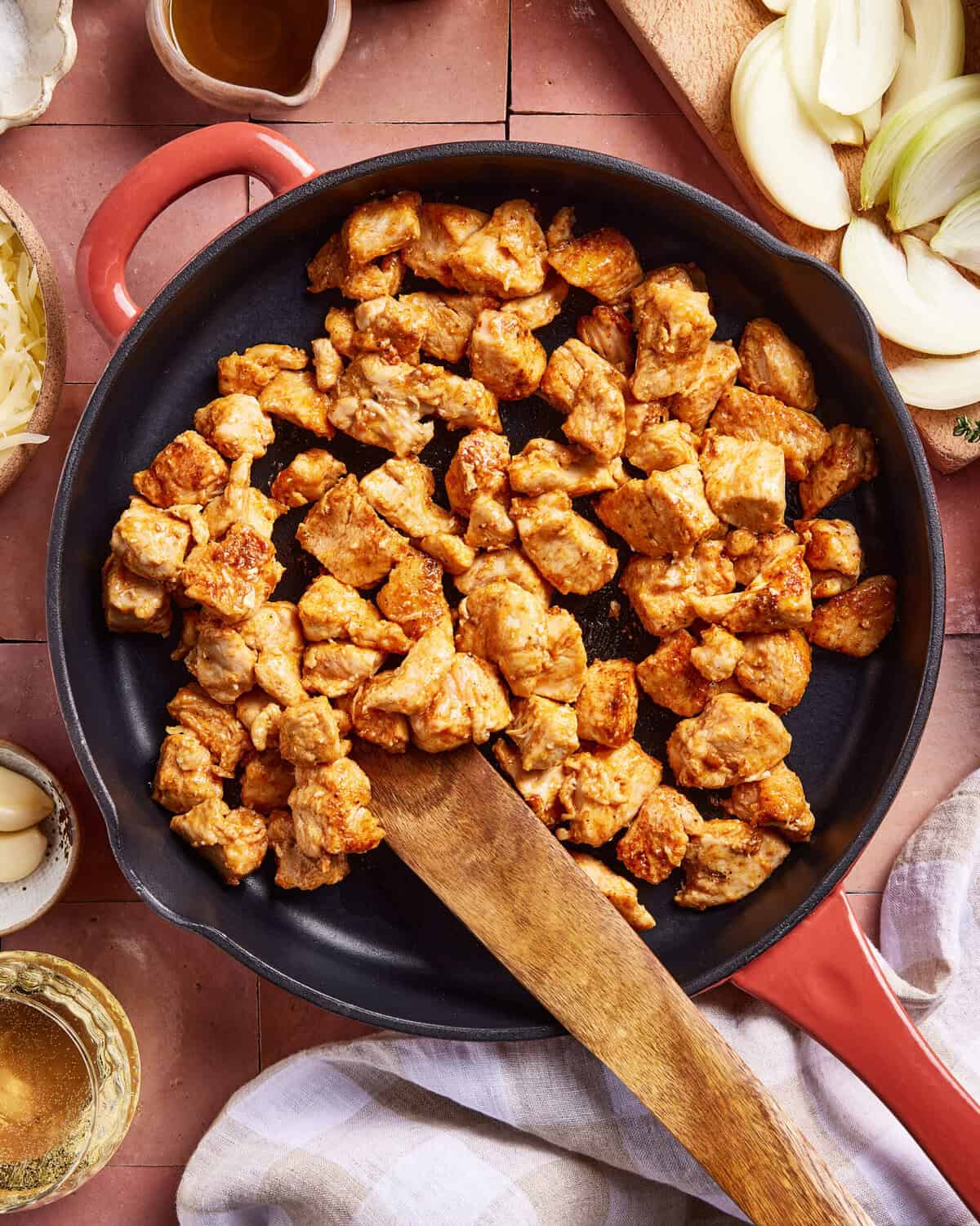 A skillet with cooked, seasoned chicken pieces being stirred by a wooden spatula. Sliced onions, garlic cloves, and small bowls of ingredients surround the pan on a tiled surface.