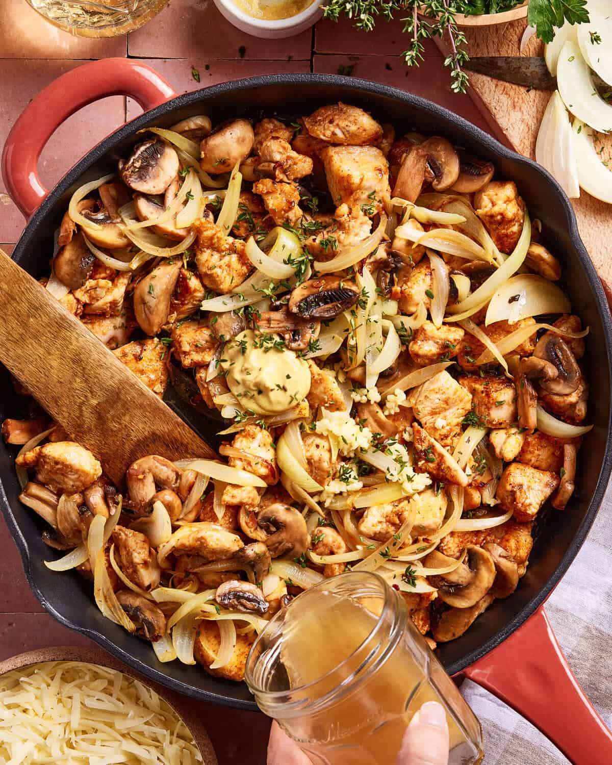 A skillet filled with cooked chicken pieces, sliced onions, and mushrooms is being stirred with a wooden spatula. A hand is pouring liquid—possibly broth—into the pan, surrounded by fresh herbs and cheese.