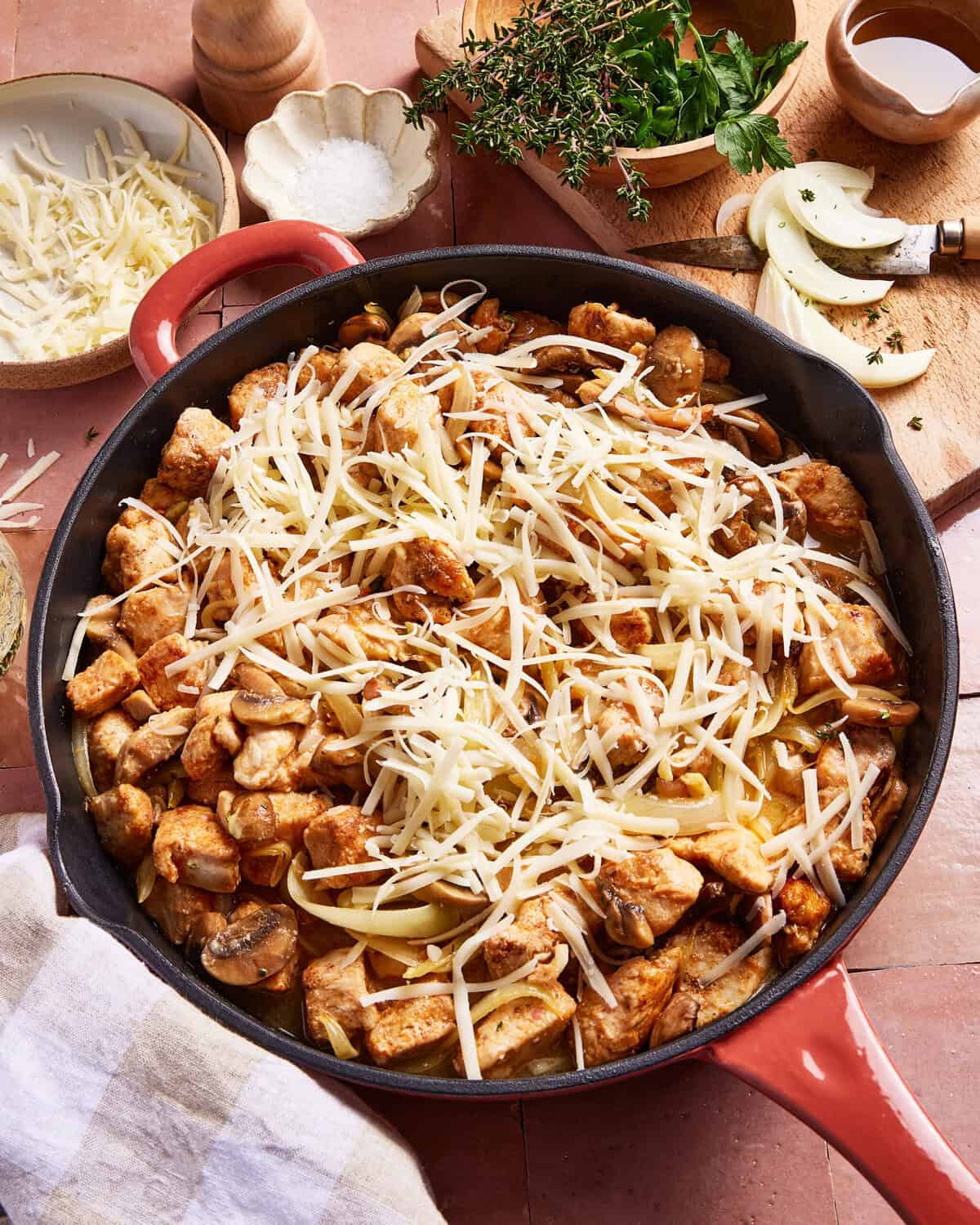 A skillet filled with cooked chicken pieces, mushrooms, and sliced onions, topped with a generous layer of shredded cheese. Fresh herbs, a wooden bowl, and a striped towel are nearby on a pink tiled surface.