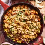 A skillet filled with cooked chicken pieces, melted cheese, mushrooms, and onions, garnished with fresh herbs. Sliced onions and herbs are on a cutting board nearby. A bowl of white rice is beside the skillet.
