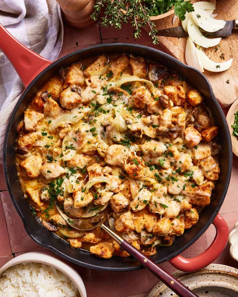 A skillet filled with cooked chicken pieces, melted cheese, mushrooms, and onions, garnished with fresh herbs. Sliced onions and herbs are on a cutting board nearby. A bowl of white rice is beside the skillet.