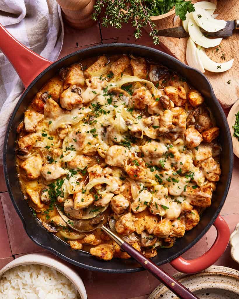 A skillet filled with cooked chicken pieces, melted cheese, mushrooms, and onions, garnished with fresh herbs. Sliced onions and herbs are on a cutting board nearby. A bowl of white rice is beside the skillet.