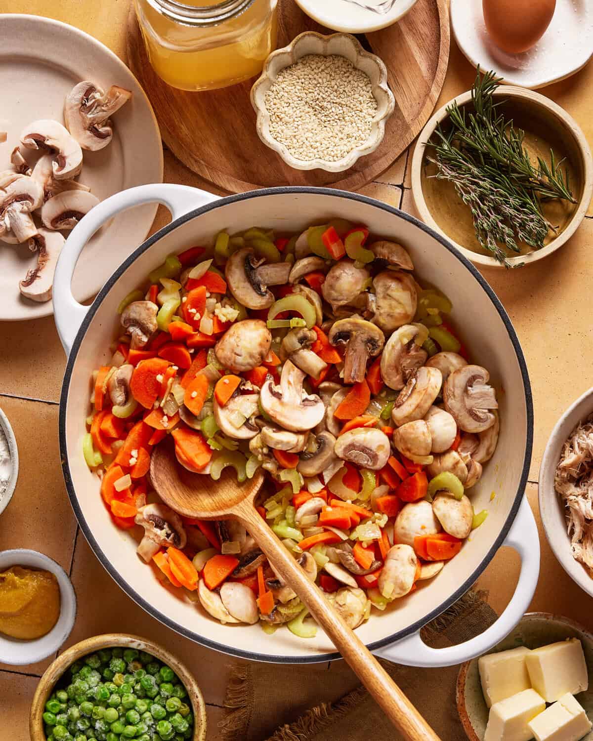 A large pot filled with sliced mushrooms, carrots, and celery being stirred with a wooden spoon, surrounded by ingredients like peas, butter, broth, herbs, and grains on a kitchen counter.