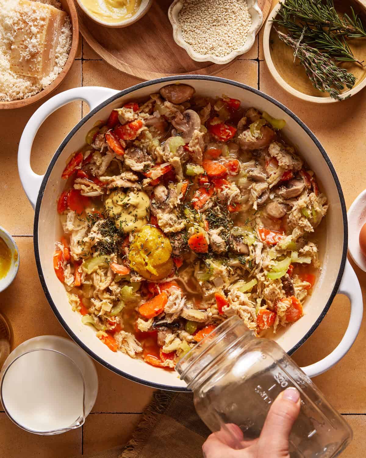 A hand pours liquid from a jar into a white pot filled with a mixture of vegetables, herbs, and shredded chicken. Surrounding the pot are bowls with ingredients like cheese, grains, and fresh rosemary.