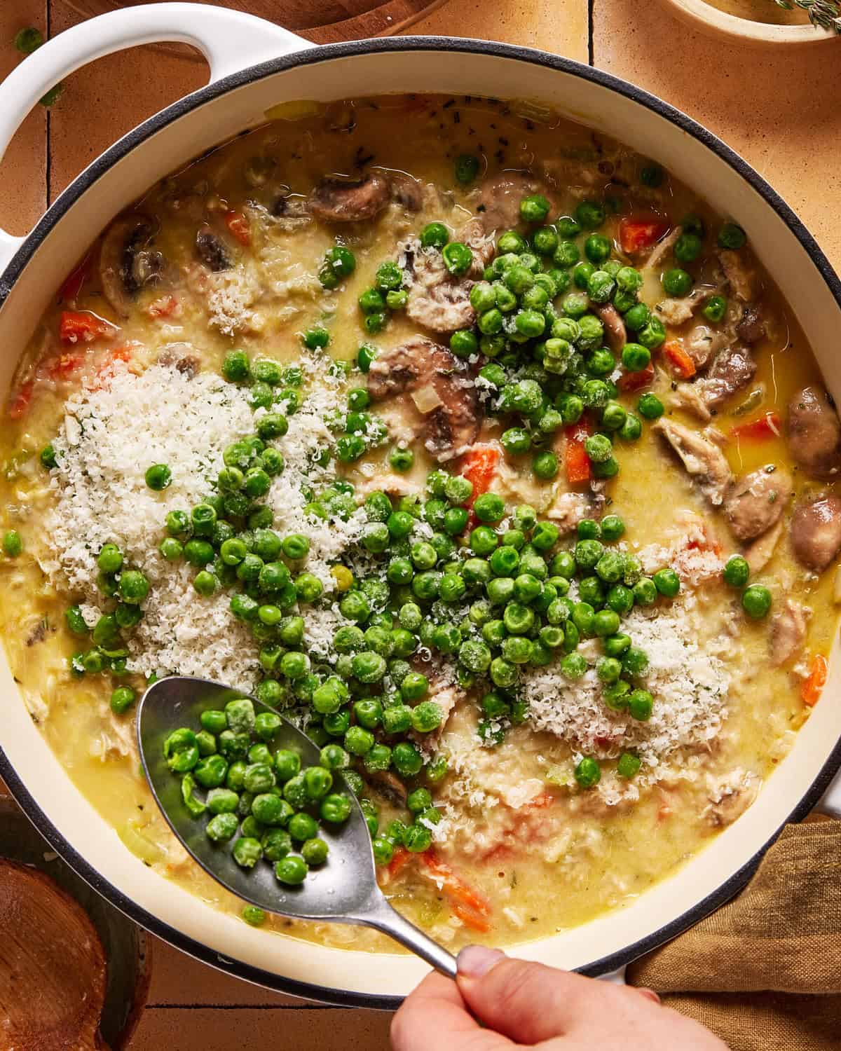 A hand holds a spoon, adding green peas to a creamy chicken and vegetable dish in a large white pot. The dish also contains mushrooms, diced red peppers, and a sprinkle of shredded cheese.