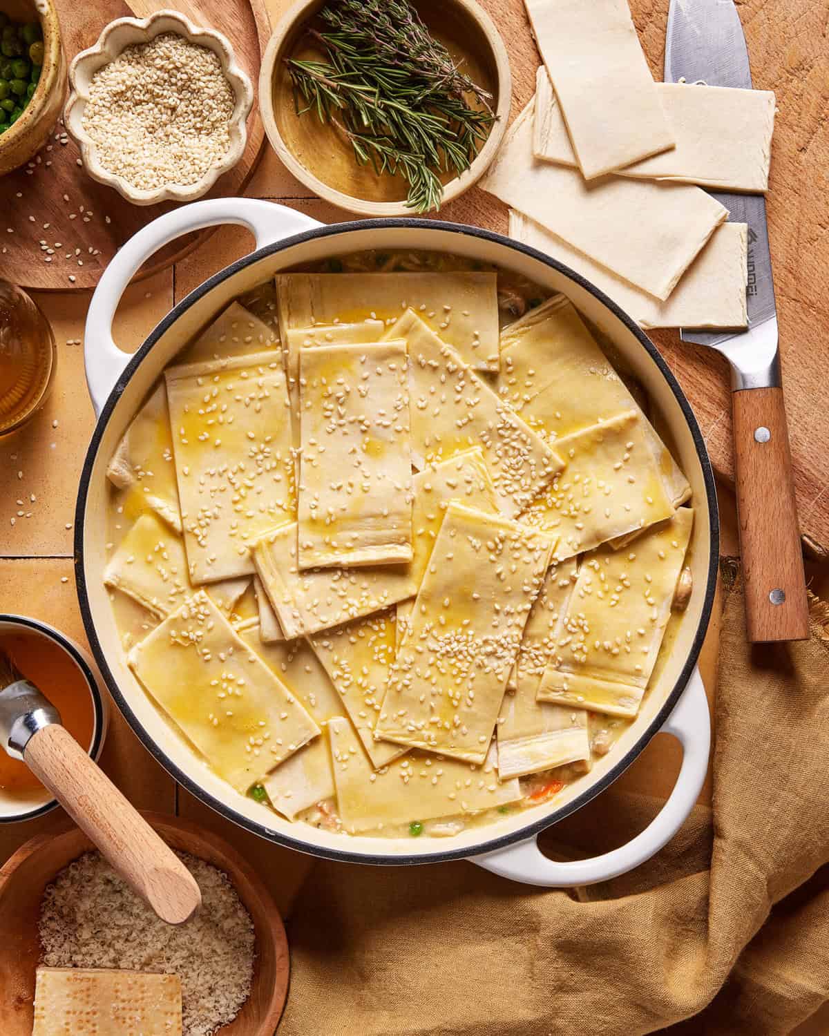 A round baking dish filled with a creamy mixture topped with overlapping rectangular pieces of dough, sprinkled with sesame seeds. Surrounding the dish are herbs, sesame seeds, a knife, and wooden bowls.
