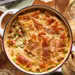 A round baking dish filled with creamy chicken pot pie topped with golden, flaky puff pastry pieces sprinkled with sesame seeds. Plates, forks, and a pepper grinder are nearby on a tiled table.