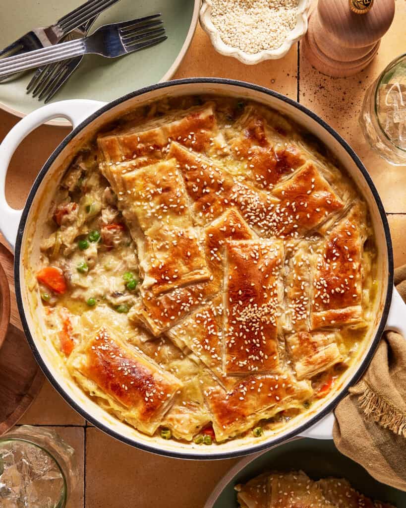 A round baking dish filled with creamy chicken pot pie topped with golden, flaky puff pastry pieces sprinkled with sesame seeds. Plates, forks, and a pepper grinder are nearby on a tiled table.