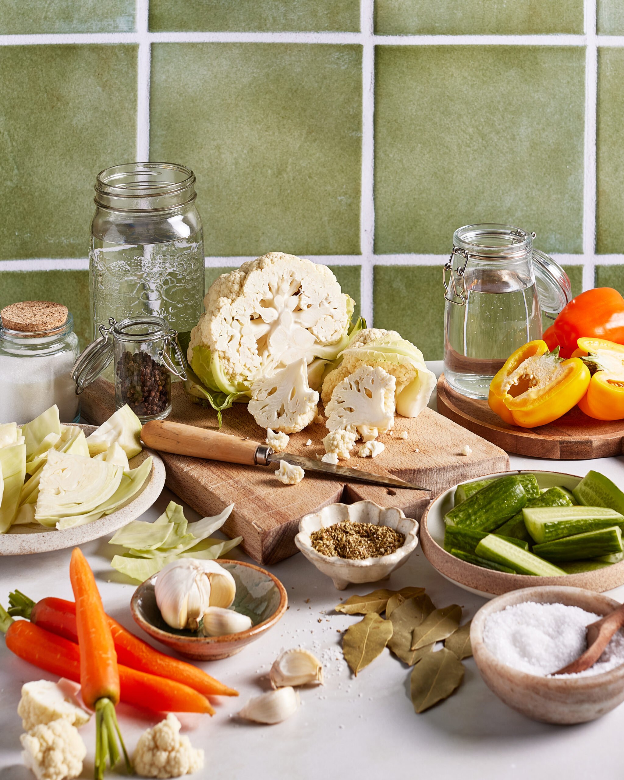 A kitchen counter with chopped cauliflower, cabbage, carrots, cucumbers, garlic, yellow bell peppers, bay leaves, spices, salt, a mason jar, and a glass of water, set against a green tiled wall.