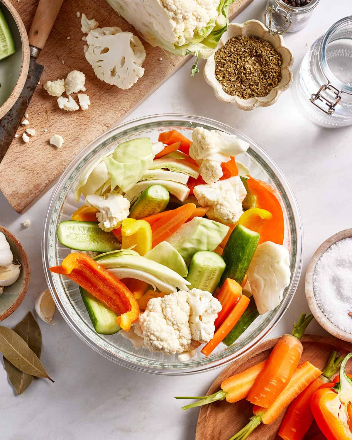 A glass bowl filled with assorted fresh vegetables, including cauliflower, carrots, cucumbers, and bell peppers, sits on a white countertop surrounded by spices, salt, and additional cut vegetables.