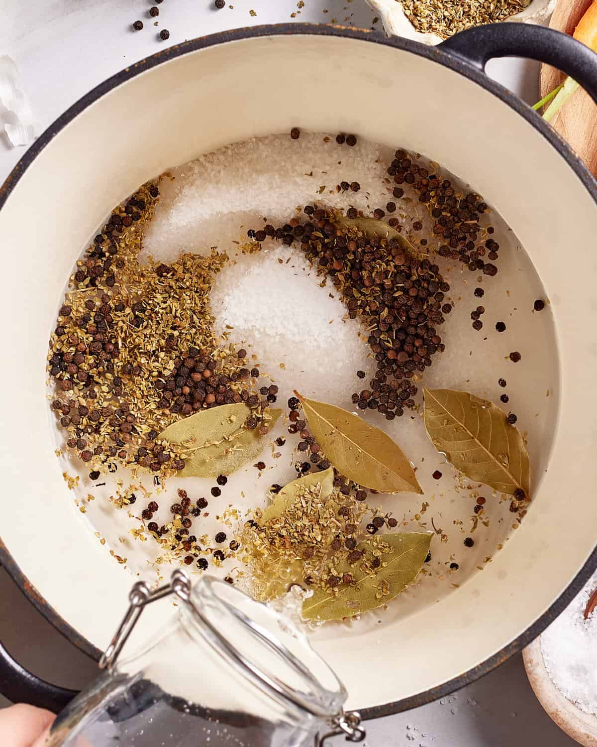 A close-up of a white pot containing water, coarse salt, whole black peppercorns, dried oregano, and bay leaves. Someone is pouring more water from a glass jar into the pot.