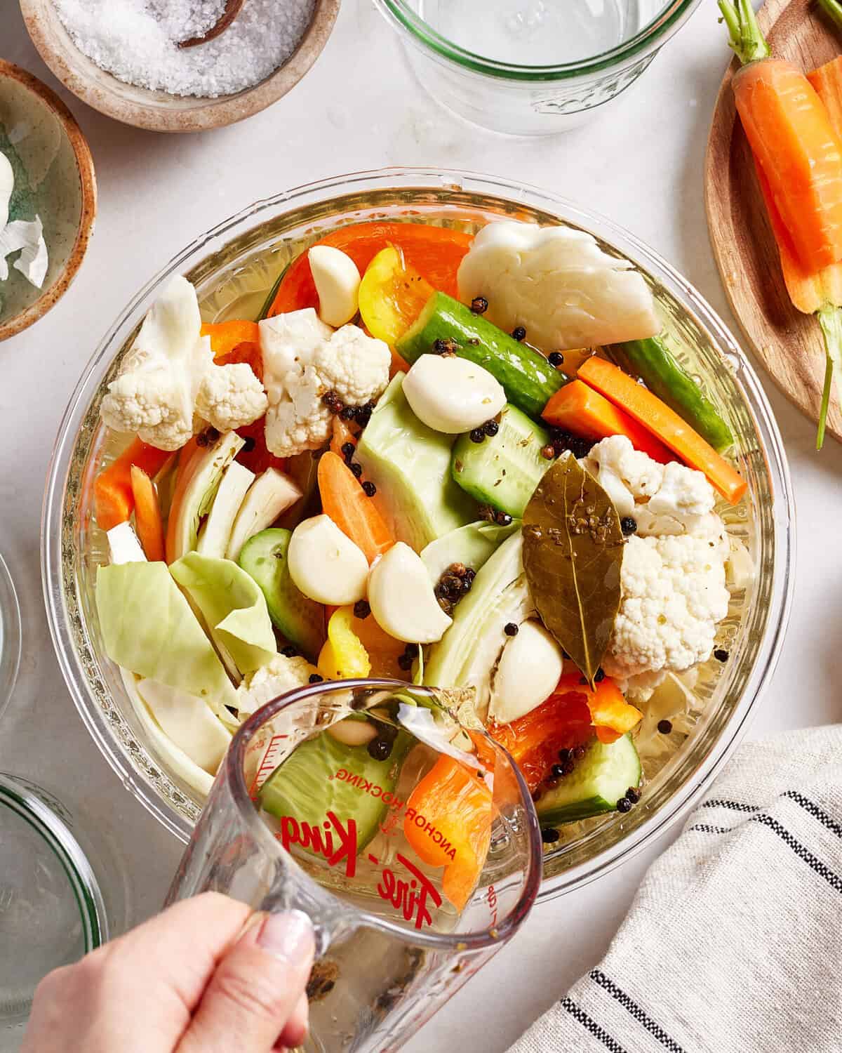 A hand pours liquid from a measuring cup into a bowl filled with sliced carrots, cucumbers, cauliflower, cabbage, garlic cloves, and spices. Other fresh vegetables and jars are visible on a white countertop.