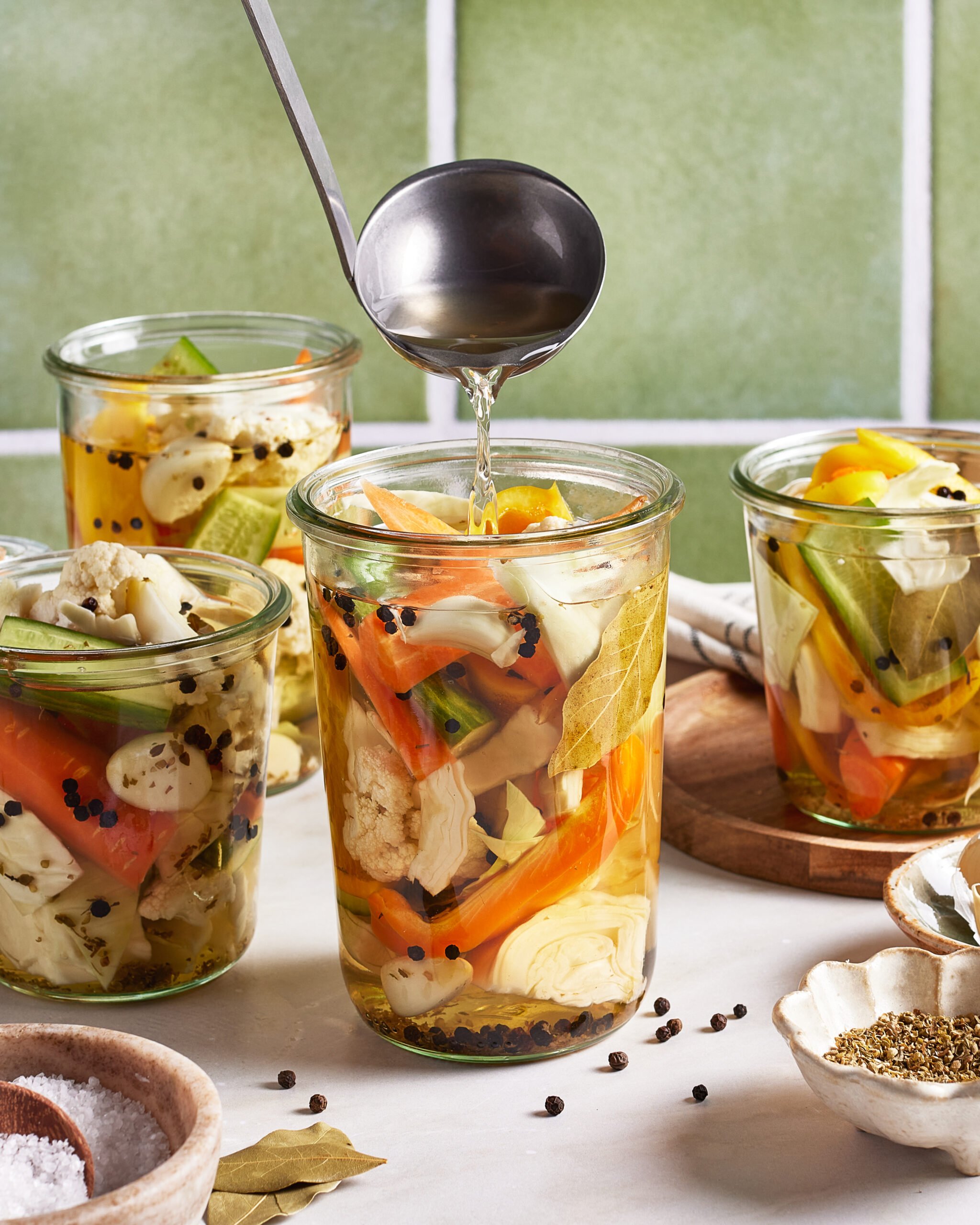 A hand pours brine from a ladle into glass jars filled with mixed vegetables, including carrots, cauliflower, and green beans, preparing homemade pickles on a kitchen counter.