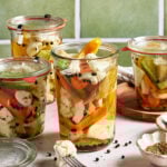 Four glass jars filled with assorted pickled vegetables, such as carrots, cauliflower, peppers, and squash, sit on a countertop with spices scattered around and green tiles in the background.