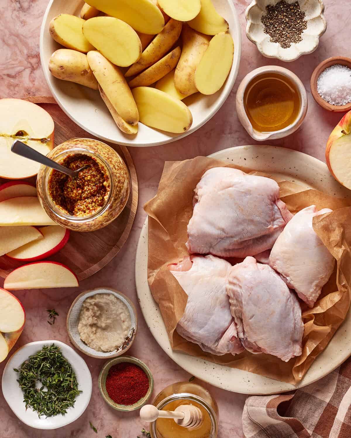 Overhead view of raw chicken thighs on parchment, sliced potatoes, apple slices, whole grain mustard, fresh herbs, ground pepper, sea salt, honey, paprika, and horseradish arranged on a pink marble surface.