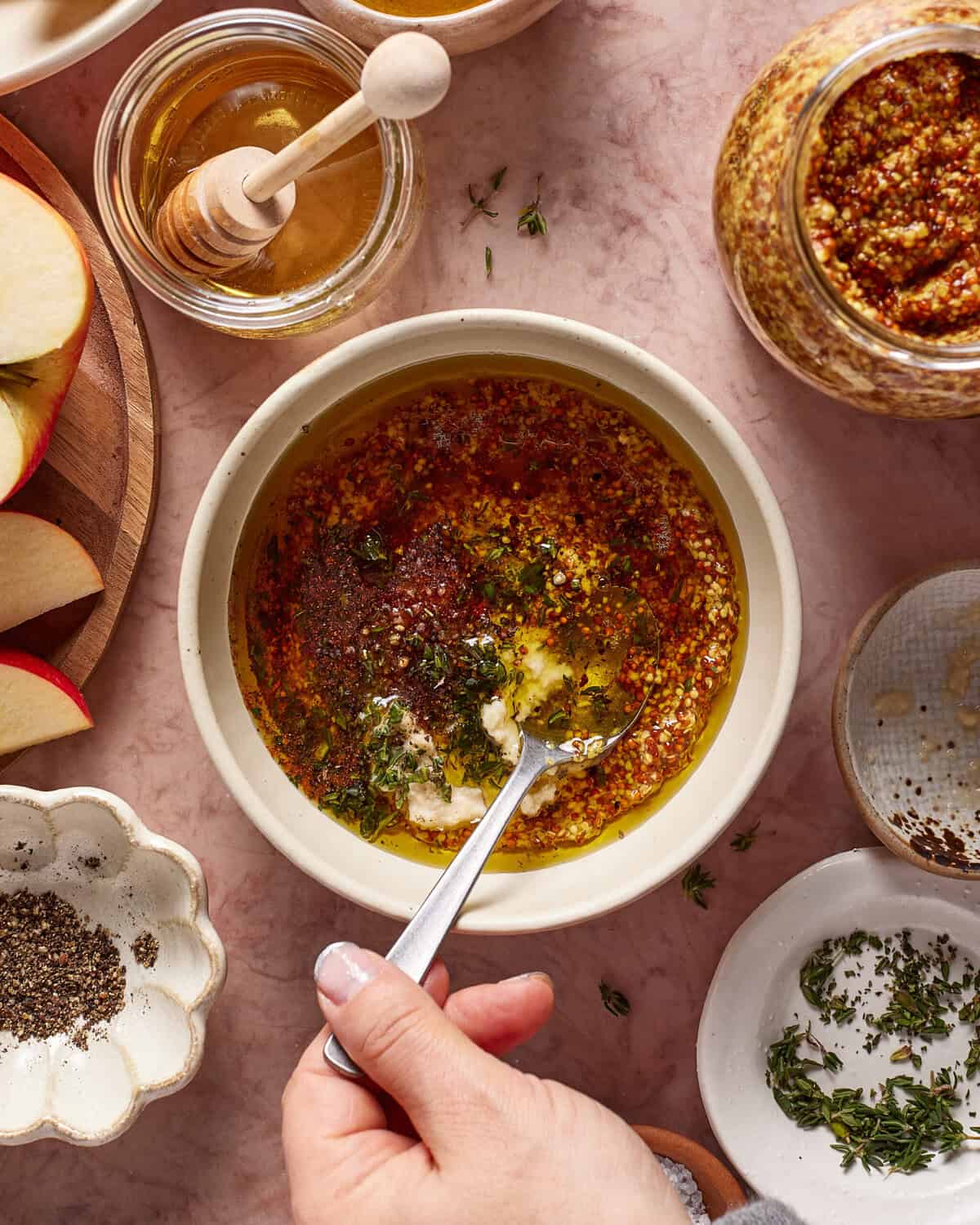A hand stirs a bowl of mustard vinaigrette surrounded by jars of honey, whole grain mustard, apple slices, black pepper, and fresh thyme on a pink marble surface.