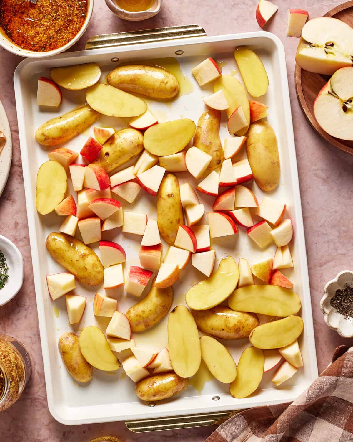 A baking tray with sliced fingerling potatoes and chopped red apples, ready to be roasted. Surrounding the tray are bowls of seasonings, sliced apples, and a checked cloth on a light countertop.