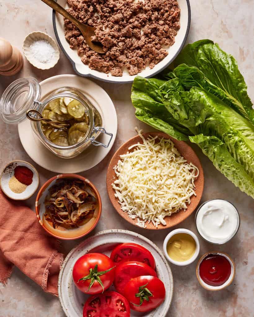 Overhead view of burger ingredients: cooked ground beef in a dish, romaine lettuce, grated cheese, sliced tomatoes, caramelized onions, pickles in a jar, small bowls of spices, ketchup, mustard, and sour cream.