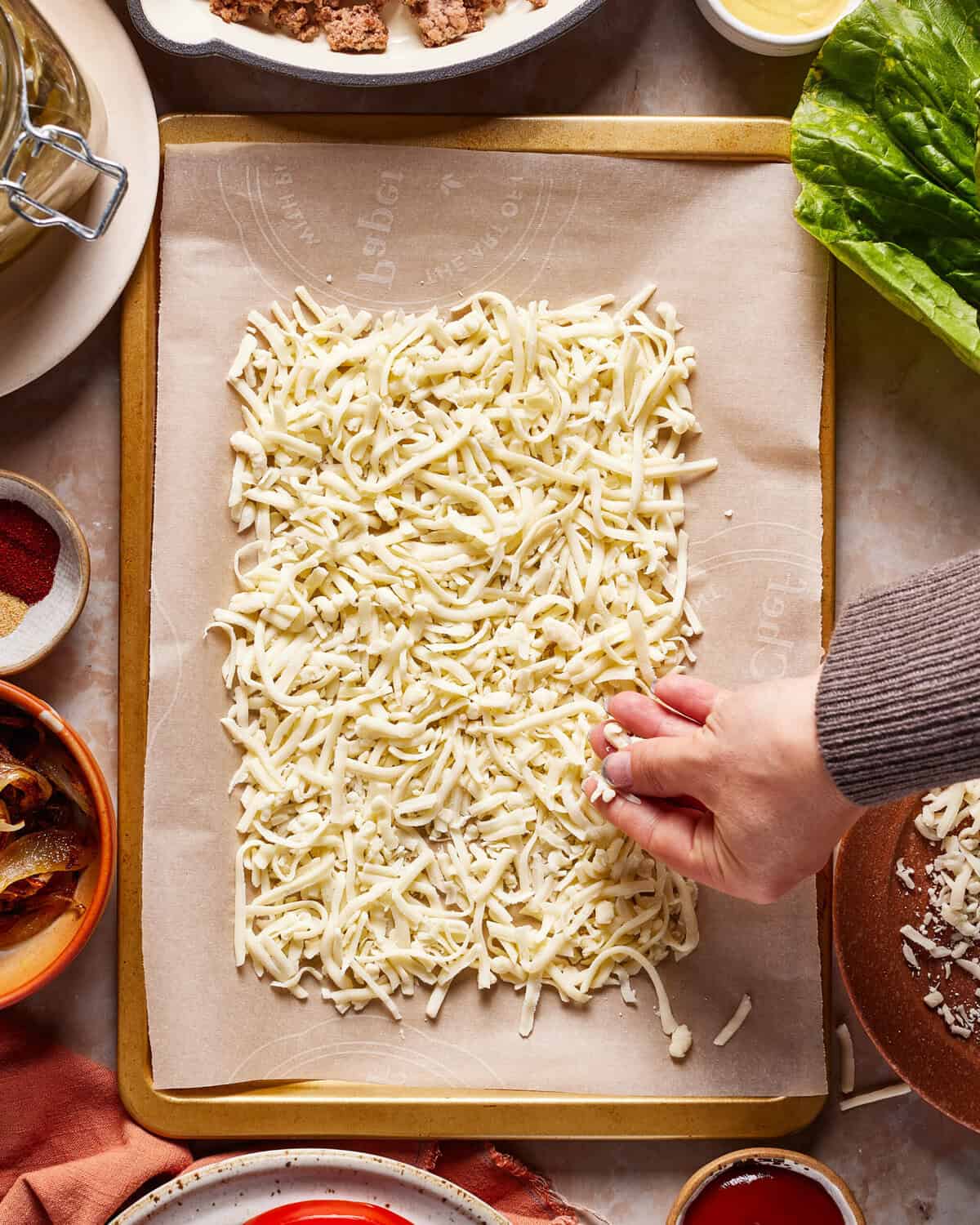 A hand sprinkles shredded mozzarella cheese onto a parchment-lined baking sheet, surrounded by various bowls of ingredients on a kitchen counter.