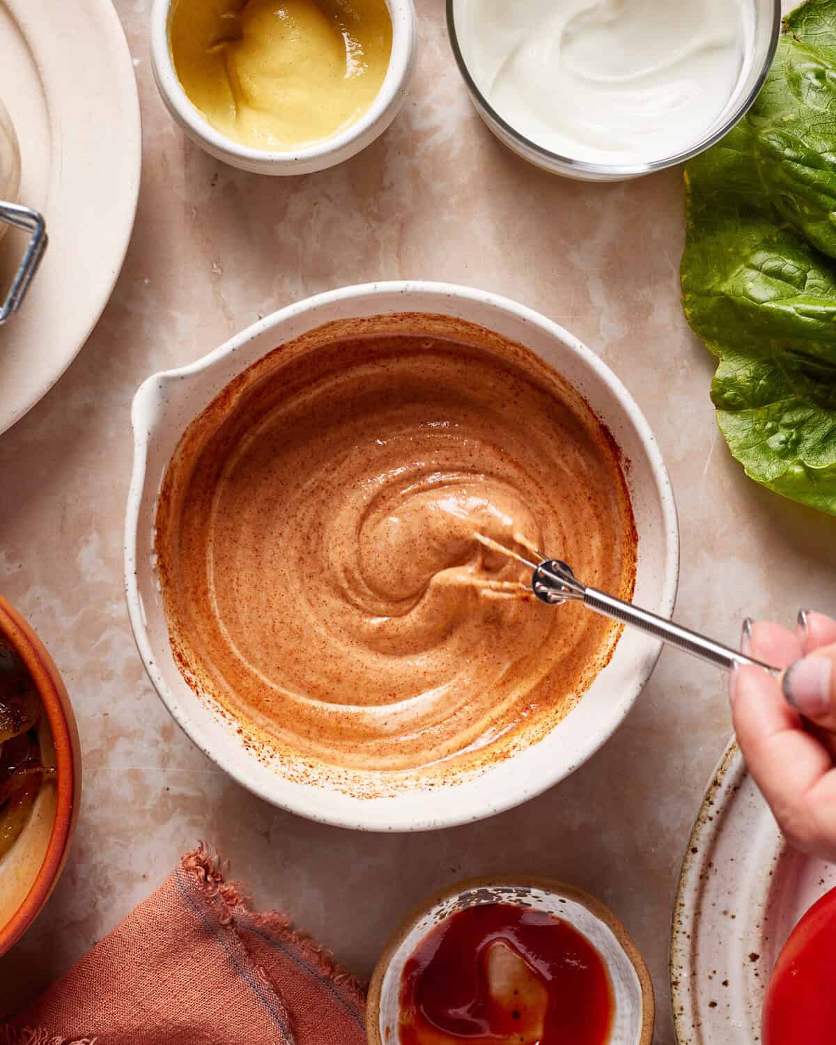 A hand stirs a creamy, reddish-brown sauce in a white bowl on a marble surface, surrounded by bowls of mustard, ketchup, mayonnaise, lettuce, and a cloth napkin.