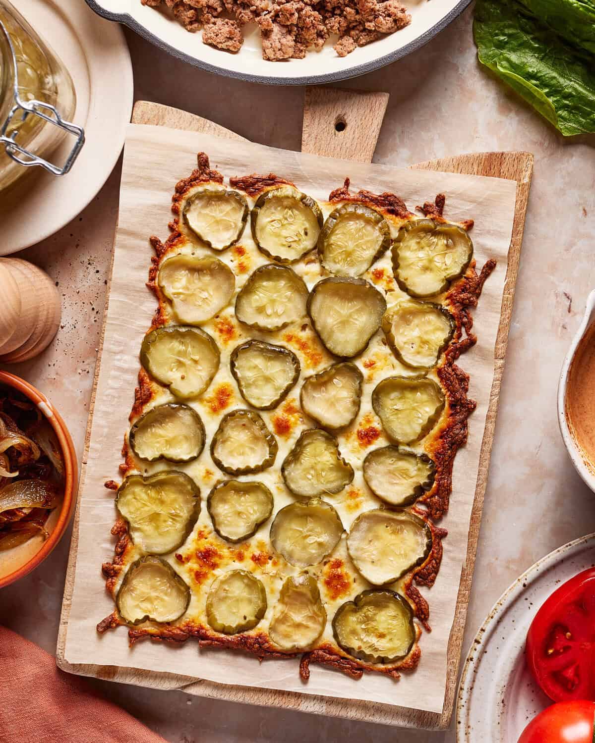 A rectangular flatbread topped with melted cheese and sliced pickles, served on a wooden board. Surrounding the board are bowls of ground meat, onions, and fresh vegetables.