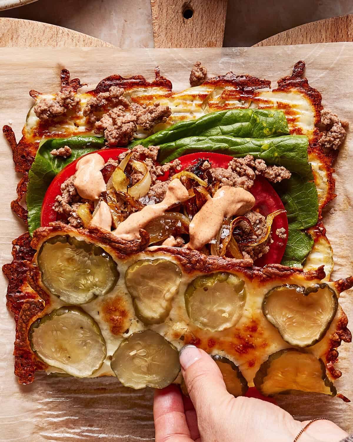 A hand lifts the edge of a cheeseburger wrap made with a melted cheese crust, topped with pickles, lettuce, tomato, ground beef, caramelized onions, and sauce, all on parchment paper.