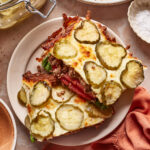 Two slices of casserole topped with melted cheese and sliced pickles are served on a plate. Ground beef, lettuce, and tomato can be seen inside. Dishes, a napkin, and jars surround the plate on a light table.