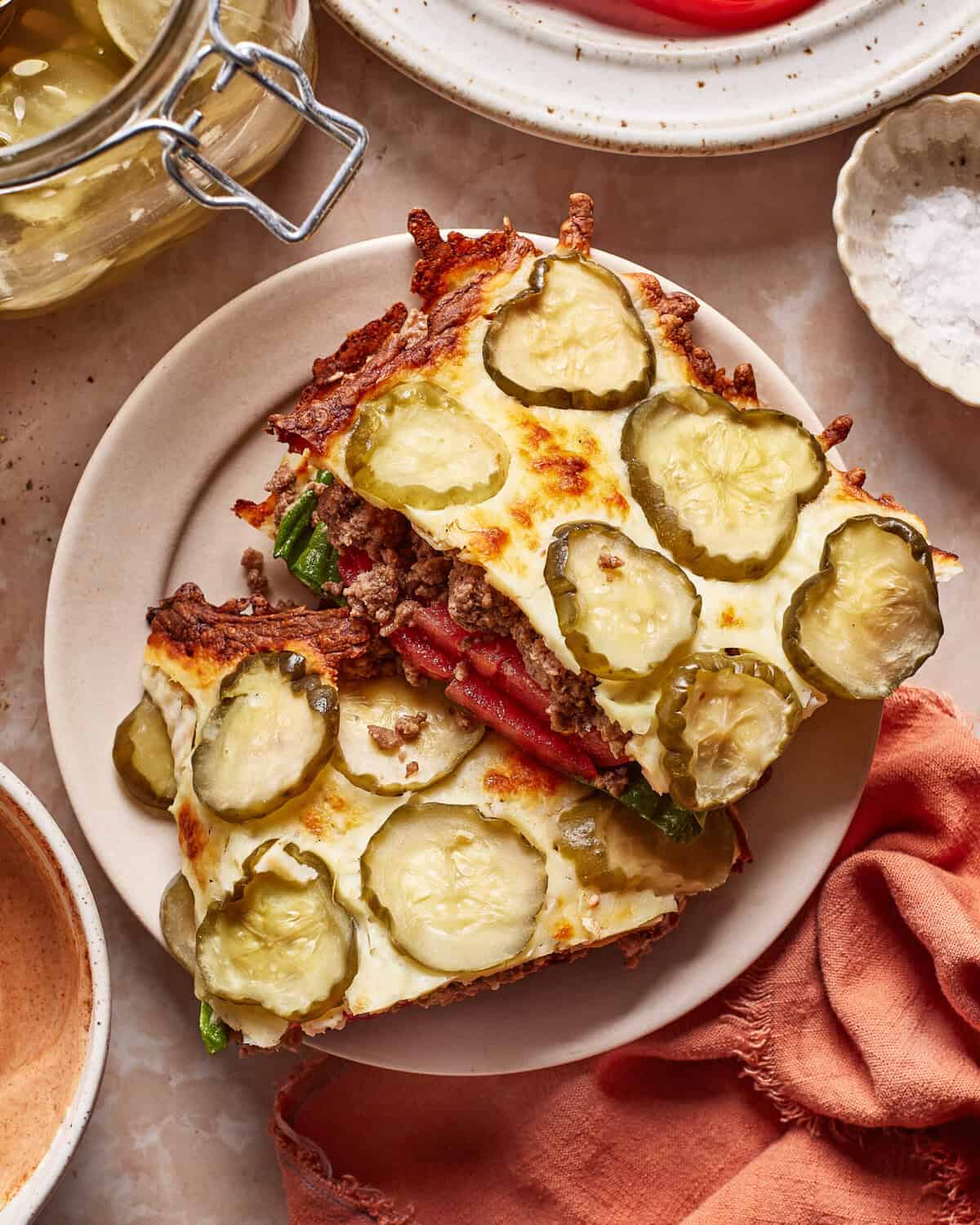Two slices of casserole topped with melted cheese and sliced pickles are served on a plate. Ground beef, lettuce, and tomato can be seen inside. Dishes, a napkin, and jars surround the plate on a light table.