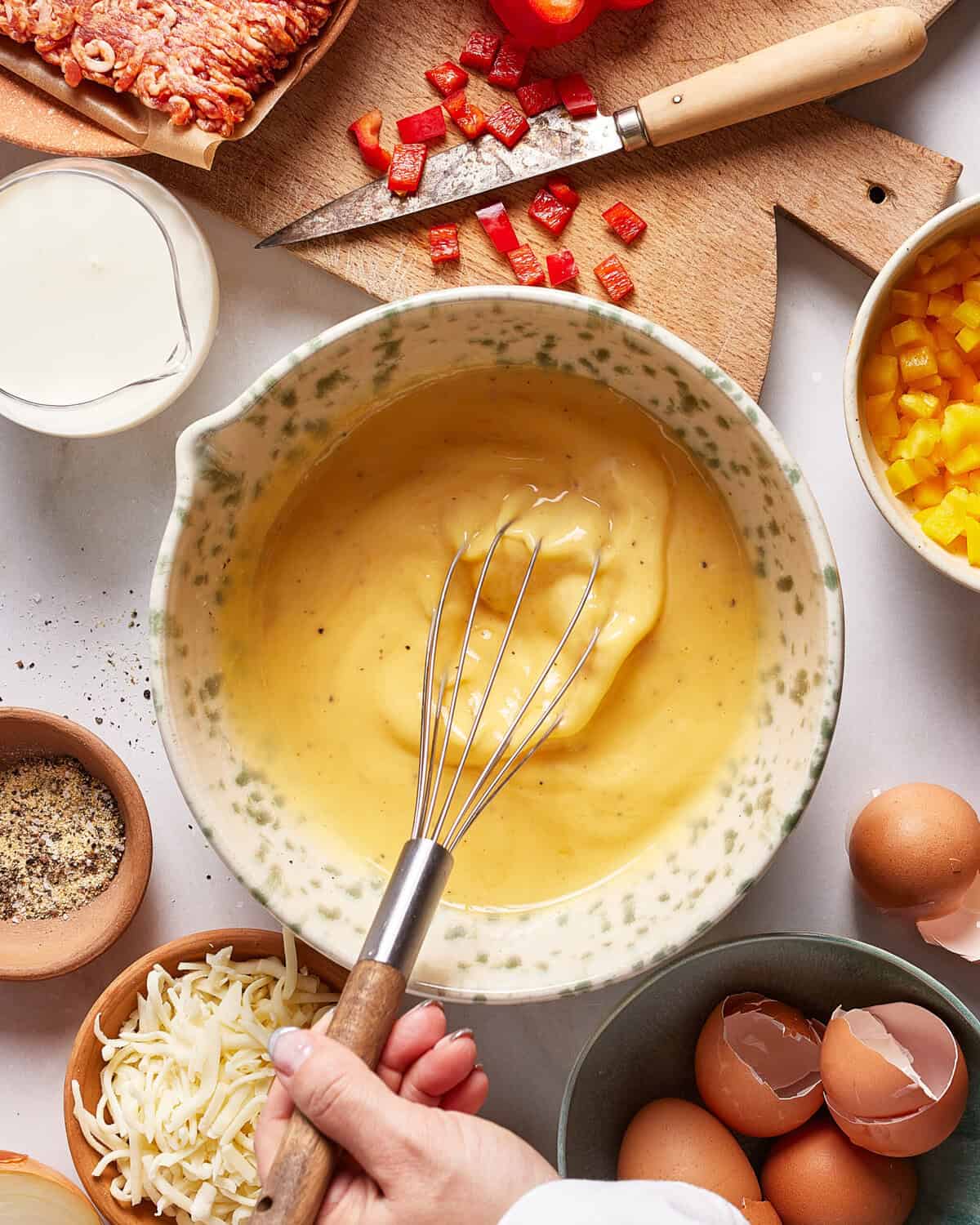 A hand whisking a yellow batter in a bowl surrounded by eggs, shredded cheese, chopped red peppers, cream, black pepper, and a wooden board with more diced peppers.