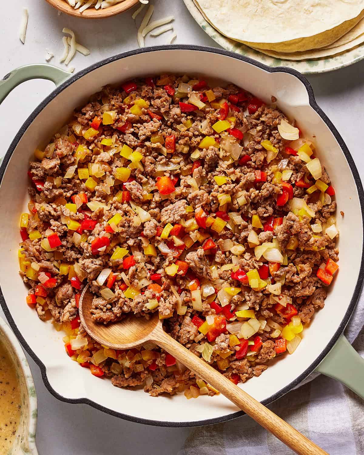 A skillet filled with cooked ground beef mixed with diced red and yellow bell peppers and onions. A wooden spoon rests in the skillet, and tortillas and shredded cheese are visible nearby.