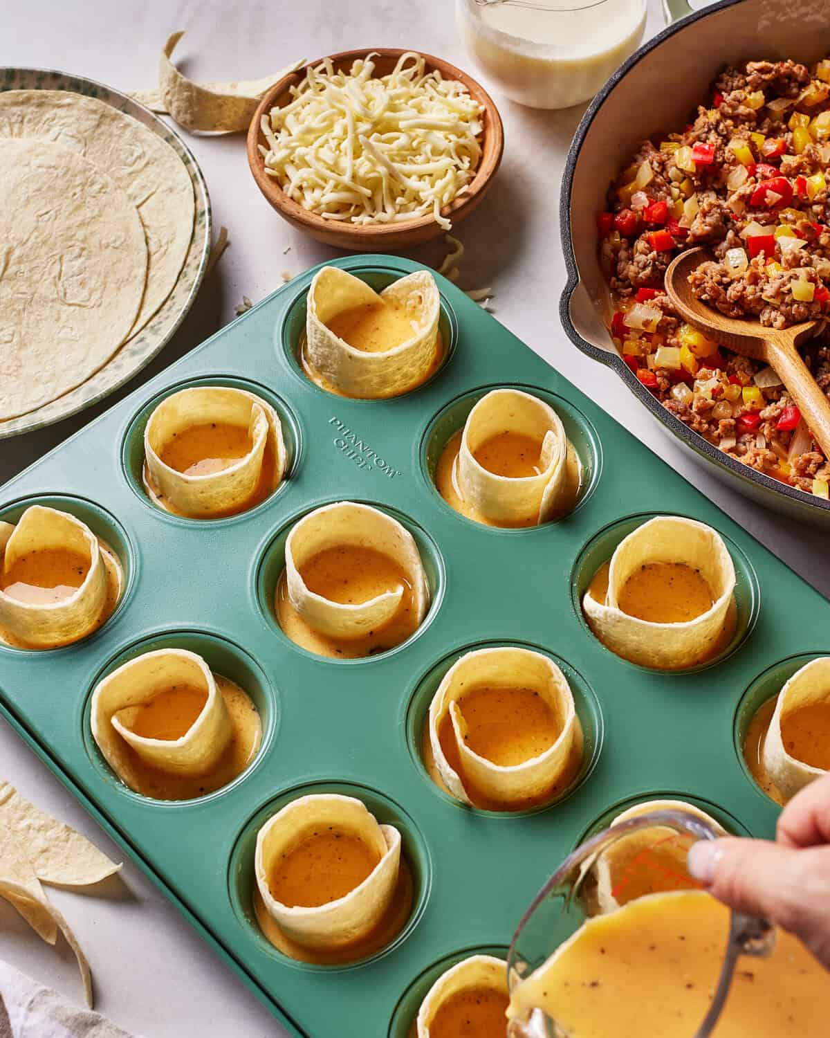 A green muffin tin filled with tortilla strips and egg mixture, surrounded by taco filling in a pot, shredded cheese in a bowl, tortillas, and a hand pouring more egg mixture into a cup.