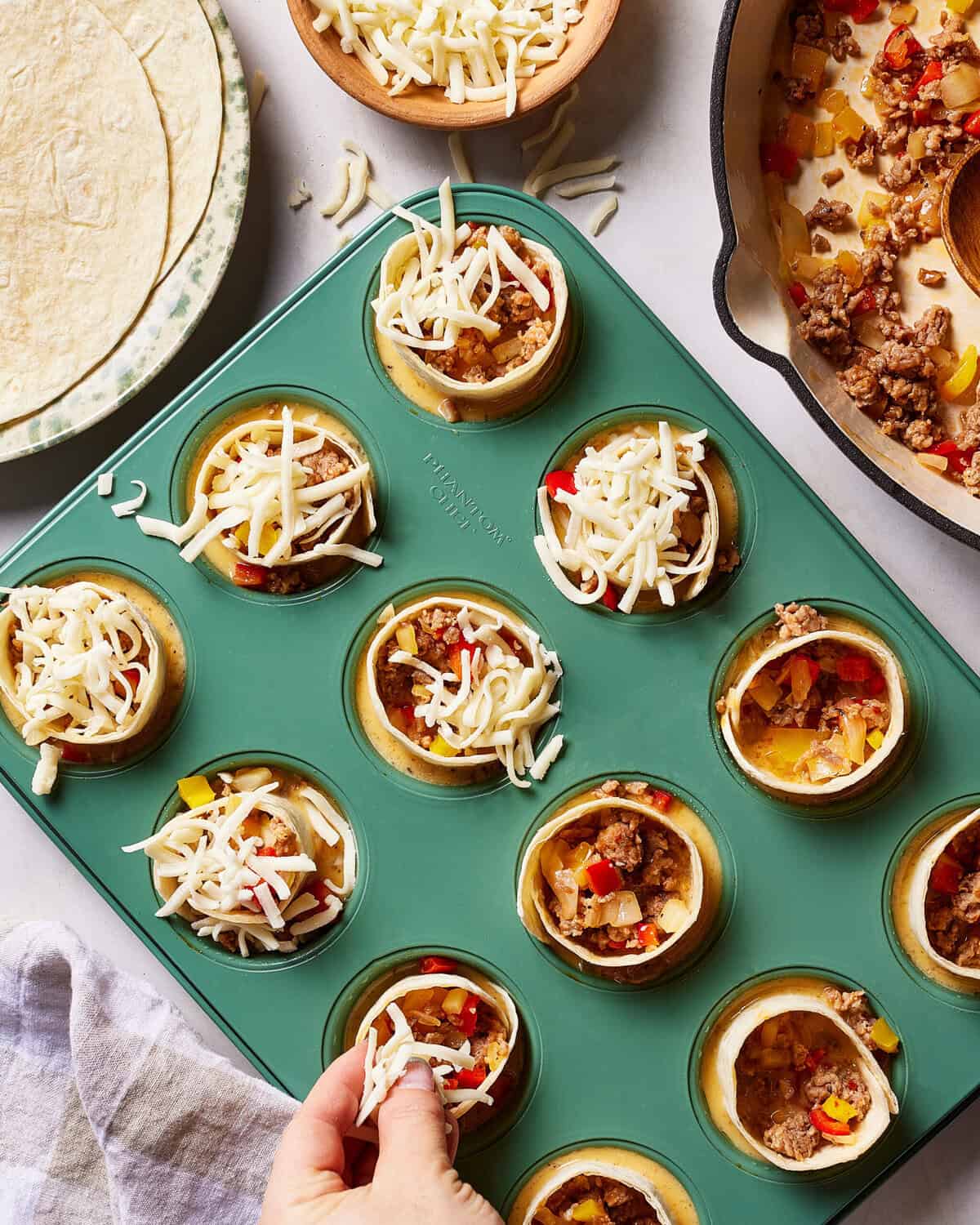 A hand sprinkles shredded cheese over mini taco cups in a muffin tin, filled with ground meat and diced vegetables. Nearby are tortillas, extra cheese, and a pot with more filling.