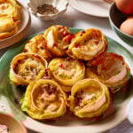 A plate of egg and cheese breakfast muffins with visible bacon pieces sits on a table surrounded by eggs, seasonings, utensils, and extra cheese.