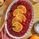 A white oval dish filled with cranberry sauce, garnished with orange slices and flaky salt, sits on a yellow surface near a bowl of salt, a pink napkin, and whole oranges and apples.