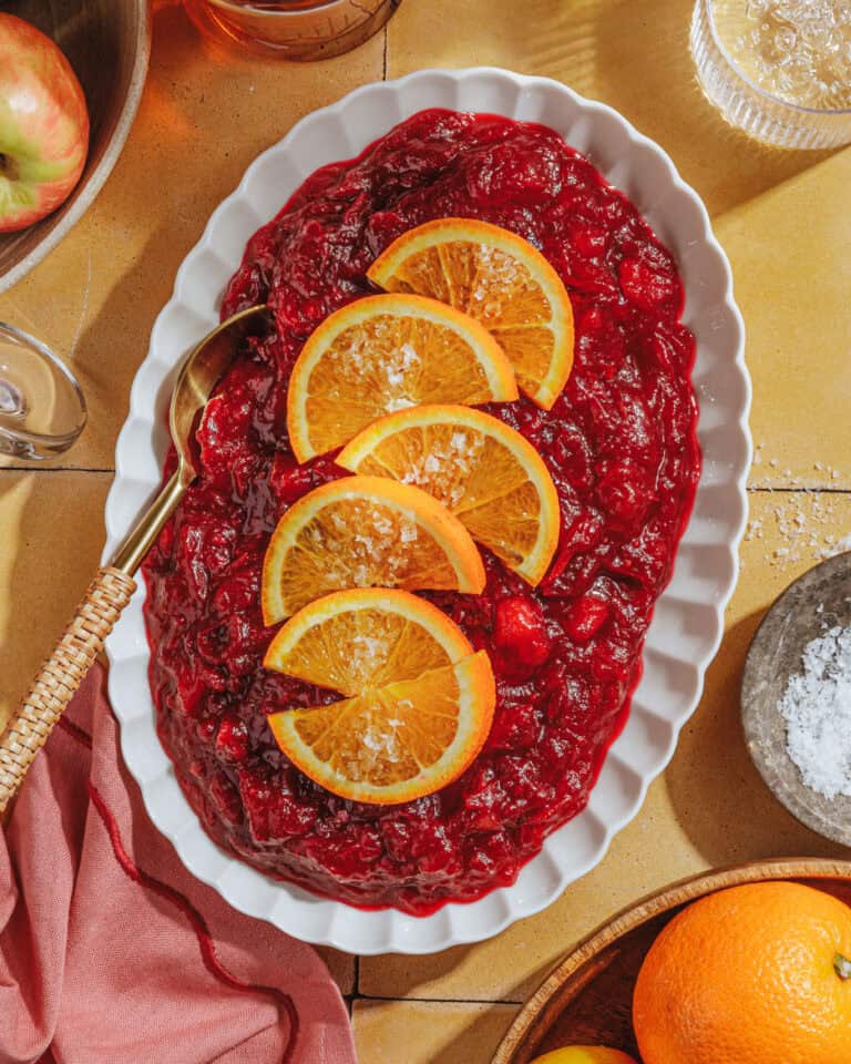 A white oval dish filled with cranberry sauce, garnished with orange slices and flaky salt, sits on a yellow surface near a bowl of salt, a pink napkin, and whole oranges and apples.