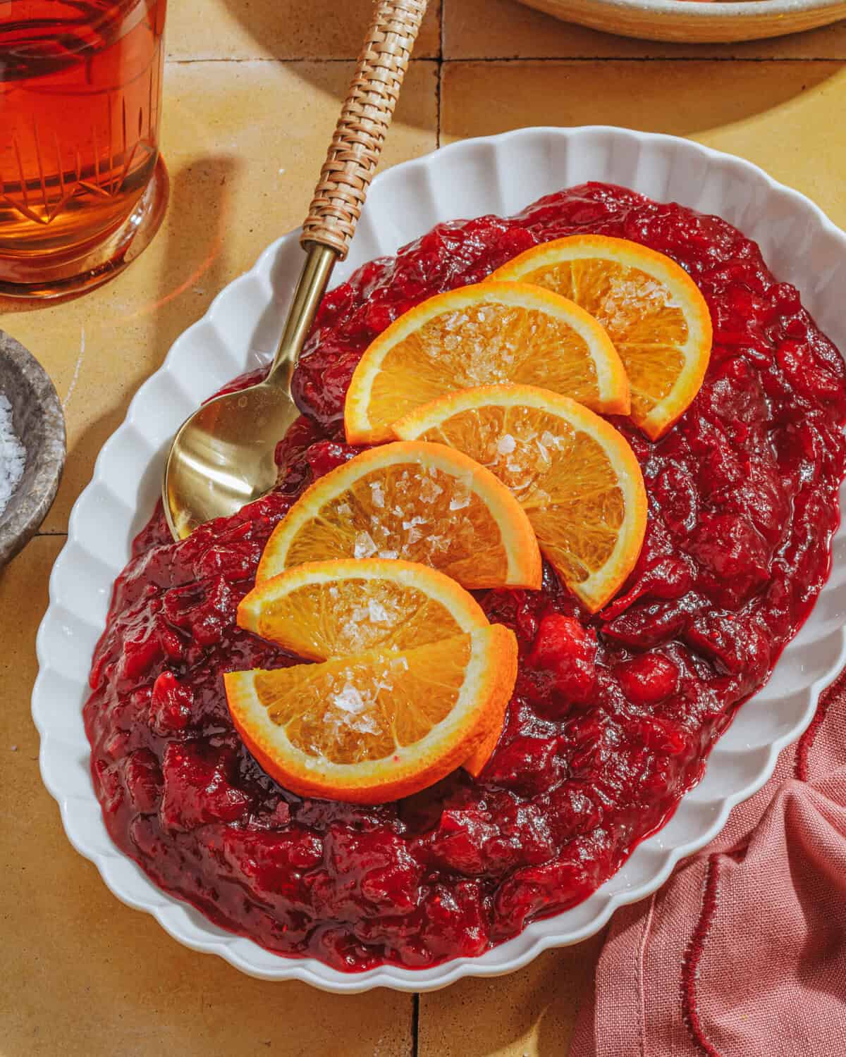 A white oval dish filled with cranberry sauce, garnished with several orange slices and a sprinkle of salt. A gold spoon with a woven handle rests in the dish. A pink napkin and a drink are nearby.