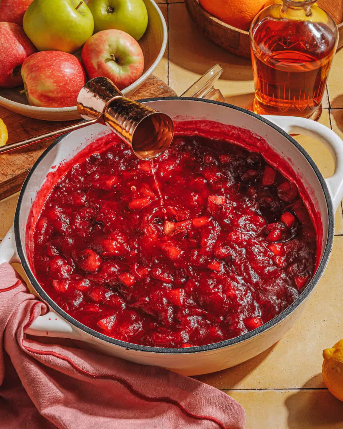 A white pot filled with chunky cranberry sauce sits on a counter. A hand pours liquid from a jigger into the sauce. Nearby are apples, a bottle of amber liquid, and a bowl of fruit. A pink cloth lies beside the pot.