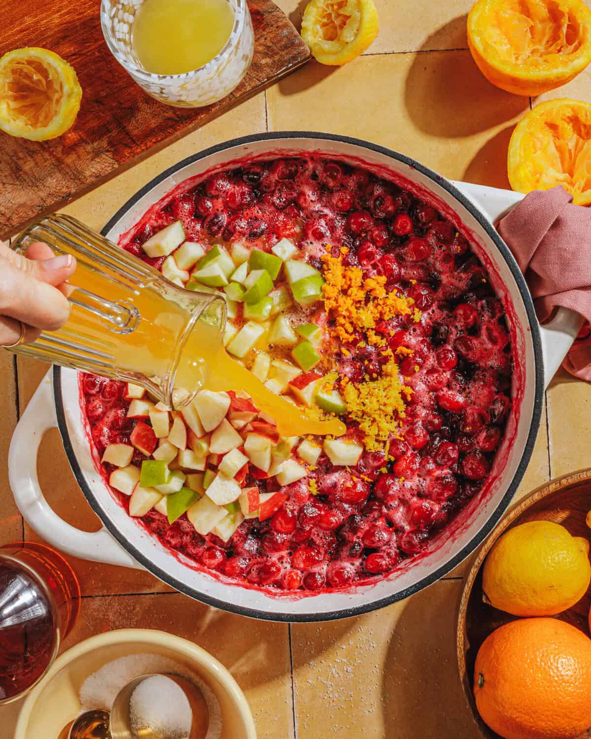 A hand pours orange juice into a pot filled with chopped apples, cranberries, and orange zest. Surrounding the pot are halved and juiced oranges, a bowl of sugar, a drink, and a small bottle on a yellow countertop.