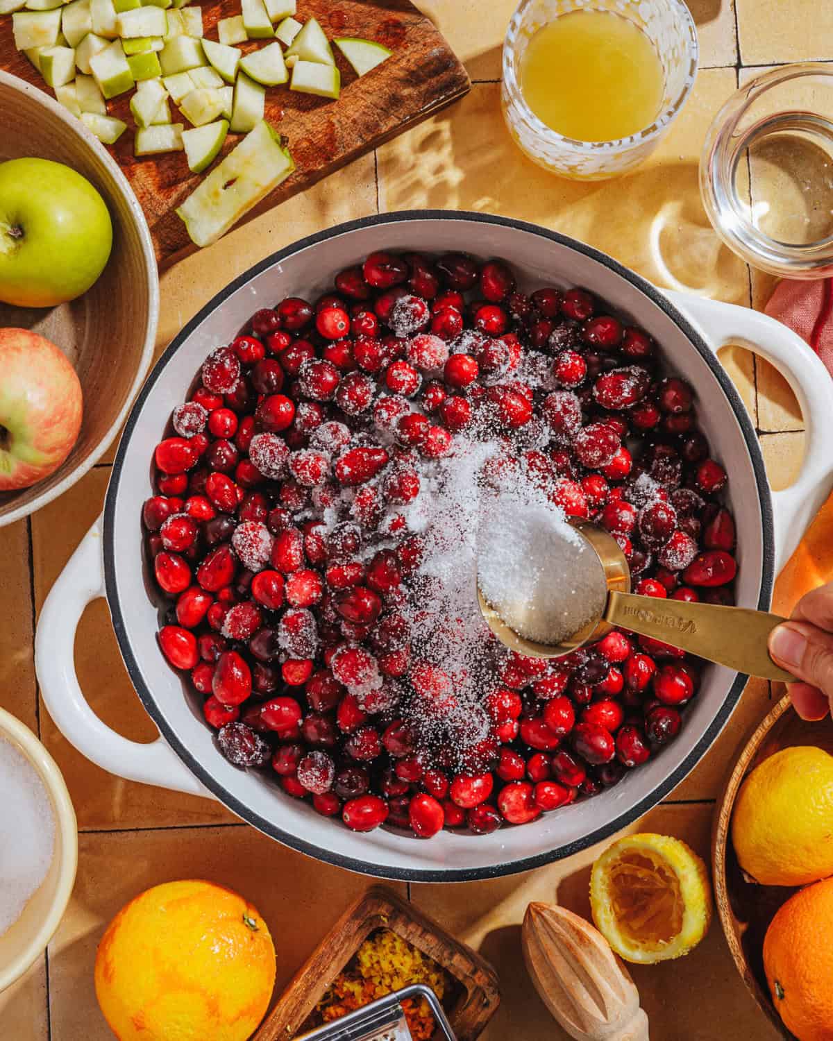 A hand sprinkles sugar over fresh cranberries in a white pot. Surrounding the pot are apples, oranges, apple pieces, juice, a zester, and a glass of liquid on a kitchen counter.