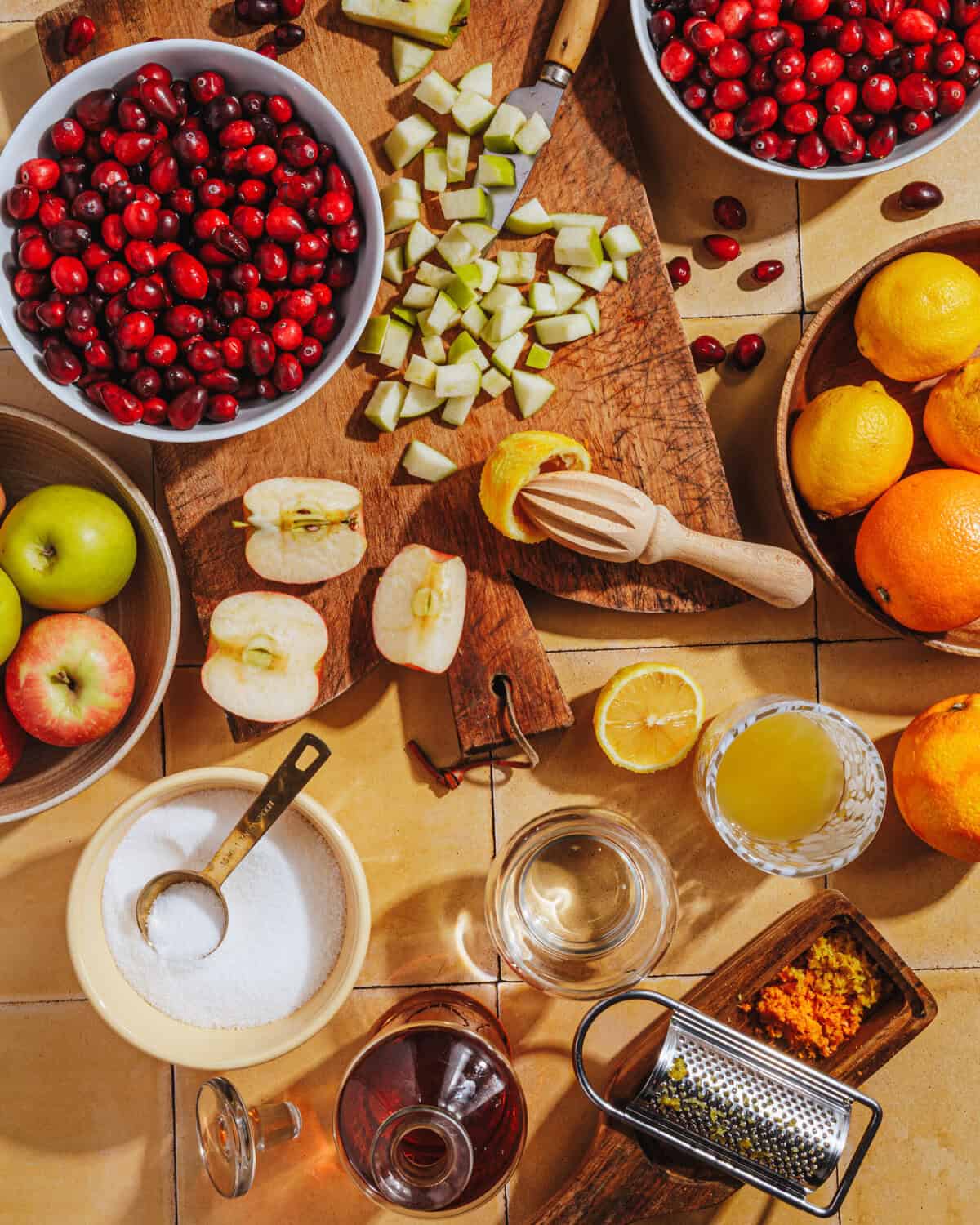 A top view of a kitchen counter with bowls of cranberries, chopped apples, whole and halved apples, citrus fruits, a juicer, a cup of sugar, a glass of juice, and zest on a grater, arranged for cooking.