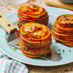 Two neatly stacked sweet potato gratin stacks garnished with chopped chives and flakes of sea salt, served on a light blue plate with a spatula. A glass of water and cutting board are in the background.