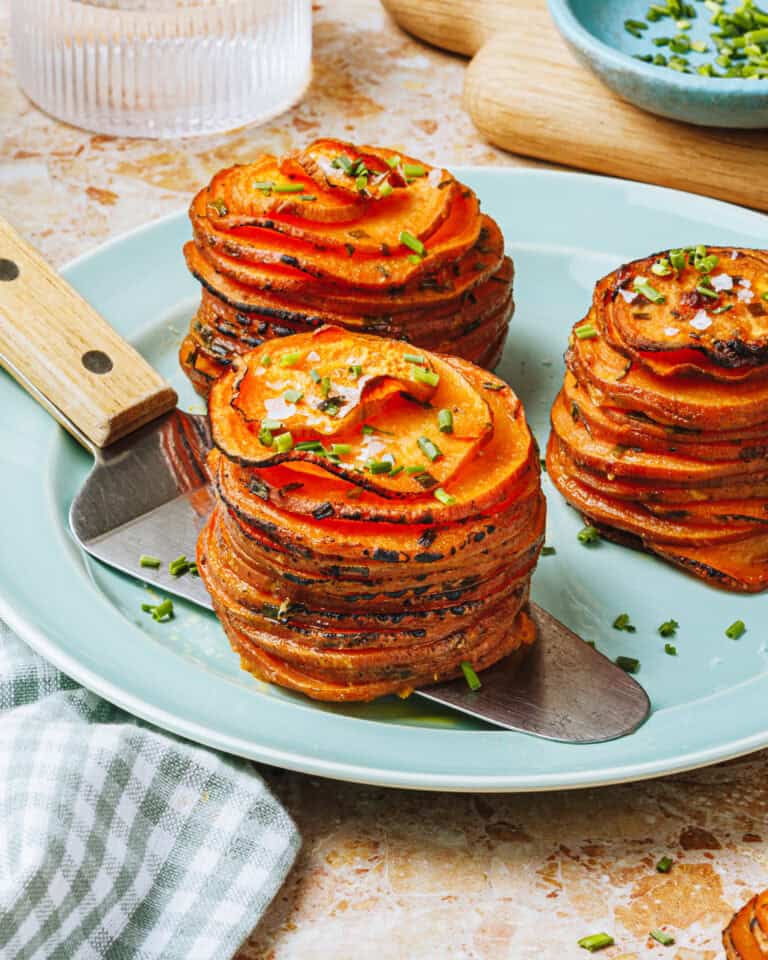 Two neatly stacked sweet potato gratin stacks garnished with chopped chives and flakes of sea salt, served on a light blue plate with a spatula. A glass of water and cutting board are in the background.