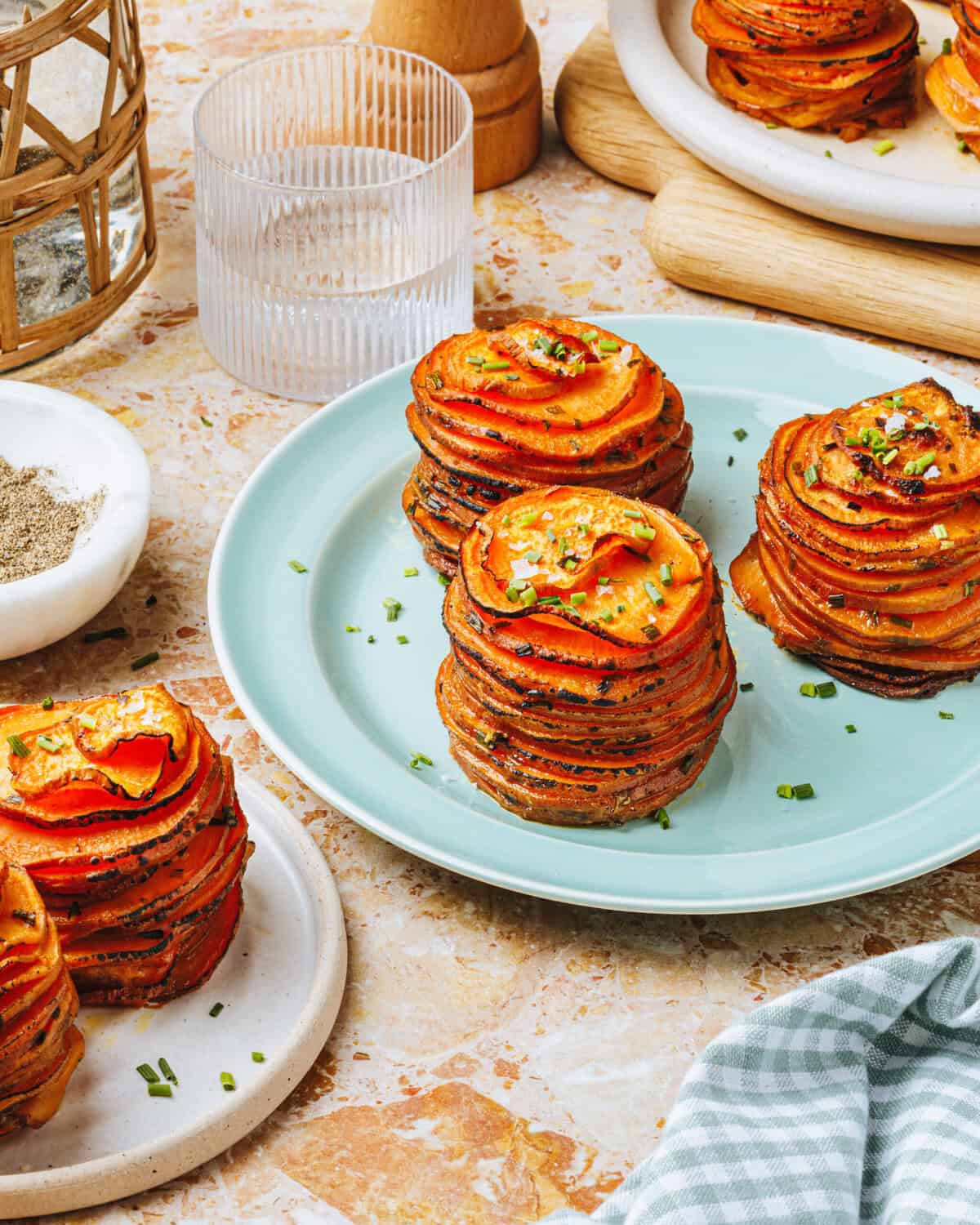 Three stacks of thinly sliced, roasted potatoes garnished with chopped chives sit on a light blue plate, surrounded by additional servings on small plates, a glass of water, and a bowl of pepper on a marble table.