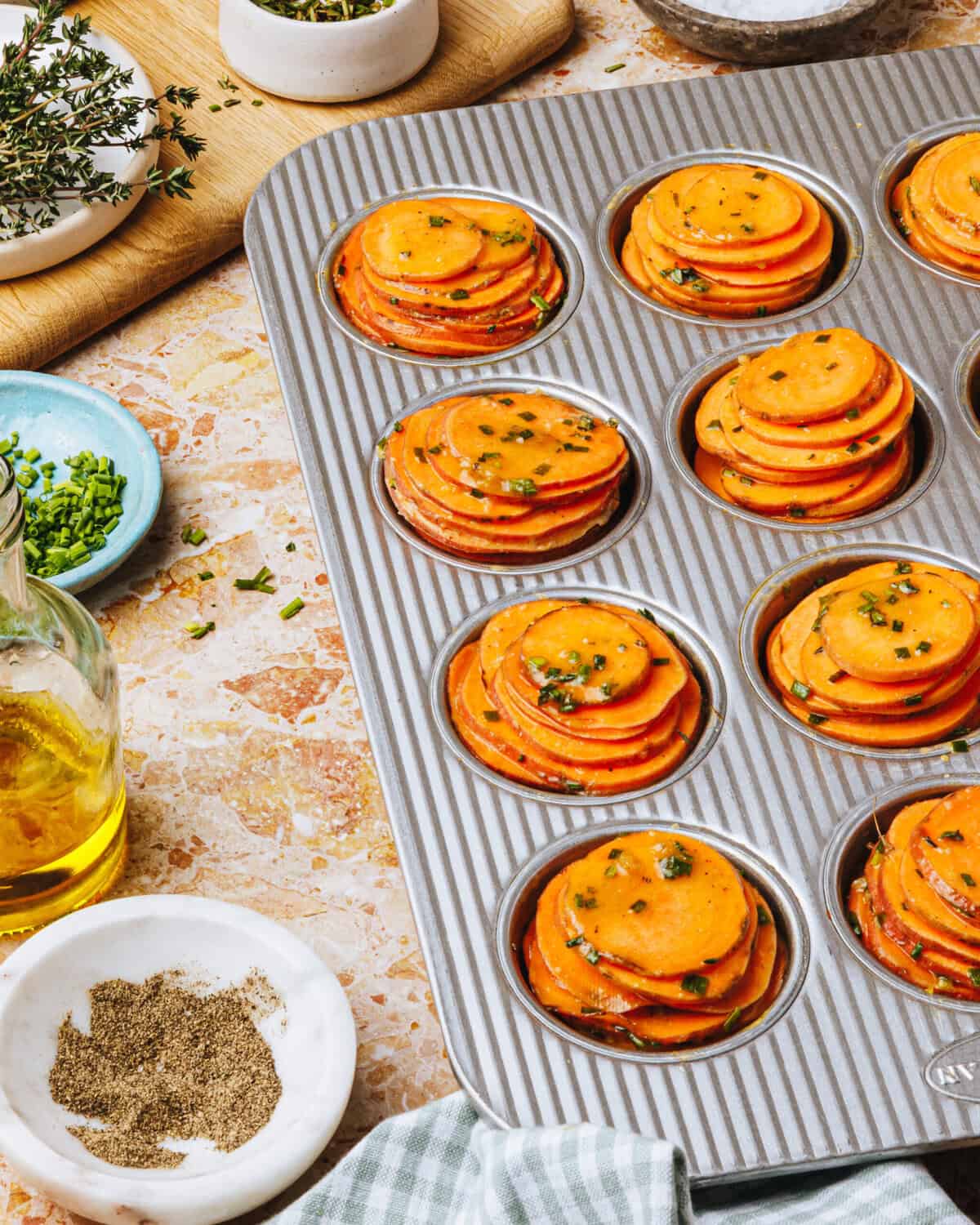 A muffin tin filled with neatly stacked sweet potato slices, garnished with herbs. Surrounding the tin are bowls of chopped chives, pepper, salt, fresh thyme, olive oil, and a cutting board with herbs.