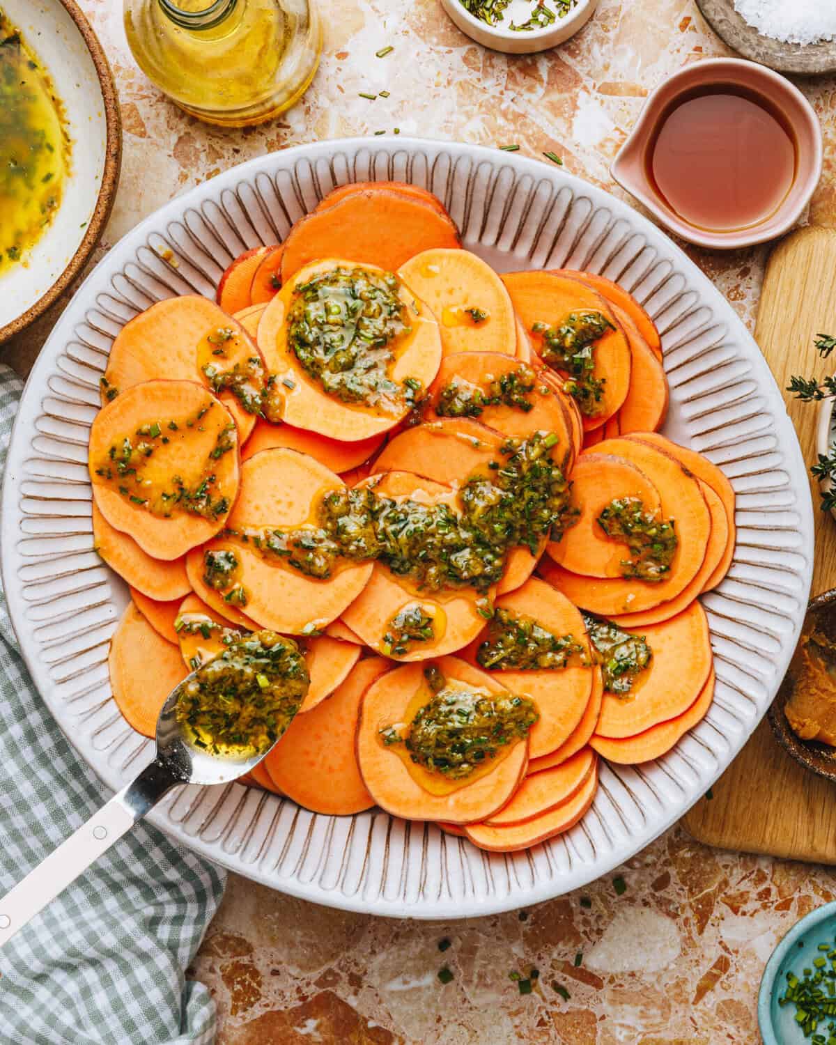 A white bowl filled with thinly sliced sweet potatoes topped with a green herb sauce. A spoon rests on the edge, and various small bowls of ingredients sit on a marble countertop nearby.